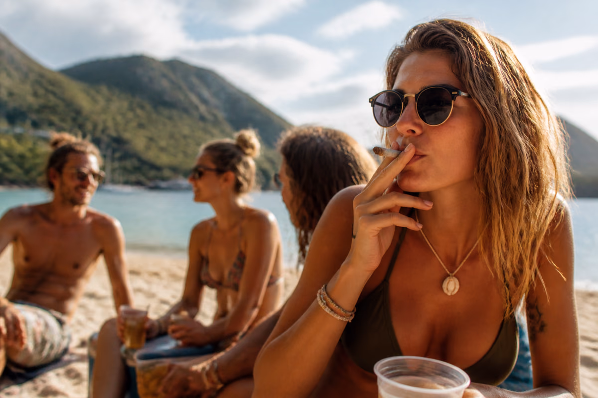 Mulher de óculos de sol fumando um baseado numa praia tropical, com amigos com bebidas ao fundo