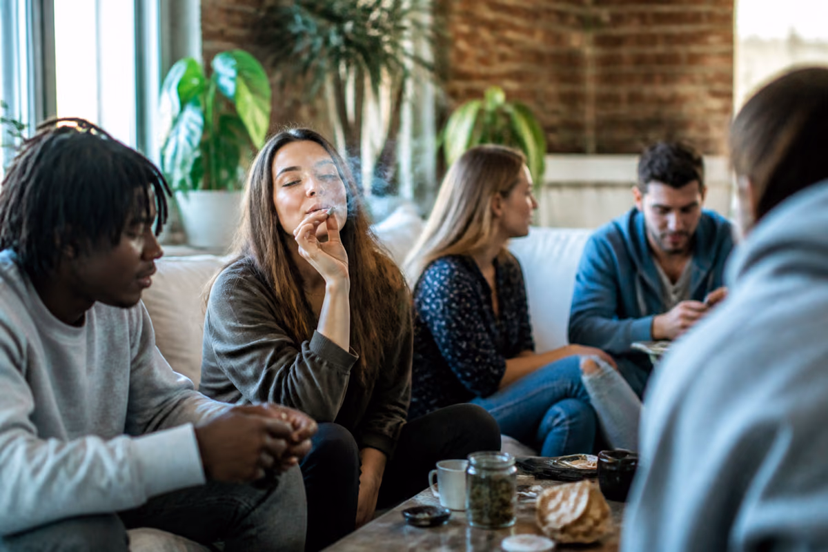 Jovens adultos num sofá, uma mulher fuma um charro, com um frasco de canábis na mesa de centro